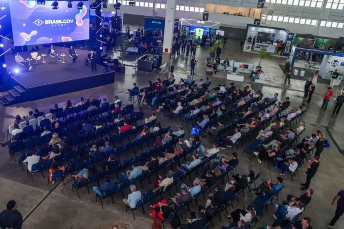 SC discute inovação pública no BrasilGov Summit 2026 Uma visão aérea panorâmica de um auditório de conferência em um grande salão de exposições. No canto superior esquerdo, um palco elevado tem um grande painel de LED exibindo "BRASILGOV SUMMIT". No palco, quatro painelistas estão sentados em poltronas modernas, e um apresentador de pé se dirige ao público. Uma grande audiência de centenas de pessoas está sentada em fileiras ordenadas de cadeiras azuis, voltadas para o palco. O fundo do salão mostra uma área de exposição com vários estandes de empresas (como "unifique" e "wacon") e pessoas circulando. A iluminação do local é natural e de palco, criando uma atmosfera ativa de evento.