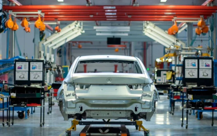 SC registra recuo de 1,5% na economia em janeiro Visão da linha de montagem de uma fábrica de automóveis moderna em Santa Catarina, mostrando a carroceria de um carro prata (BMW) sobre um carrinho de transporte industrial, cercada por equipamentos e estações de trabalho, exemplificando o setor de fabricação de veículos que registrou queda no início de 2026.