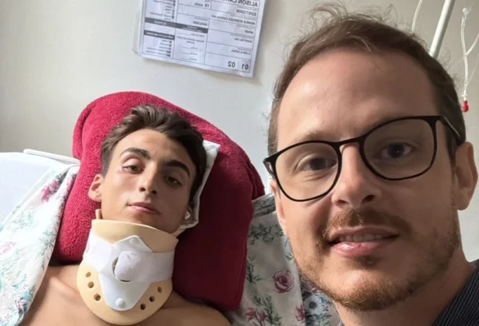 SC terá primeira aplicação de polilaminina em Sombrio Uma fotografia do tipo selfie, tirada em um quarto de hospital, mostra em primeiro plano, à direita, o Dr. Ângelo Formentin Neto, médico residente de anestesiologia e coordenador do Pronto Socorro, de óculos e sorrindo para a câmera. À esquerda, deitado na cama e apoiado por um travesseiro vermelho, está o paciente Alison Carvalho Saldívia, de 19 anos, que usa um colar cervical rígido. Alison olha diretamente para a câmera. Ao fundo, na parede branca, estão fixadas tabelas e prontuários médicos. A imagem ilustra o médico que viabilizou o tratamento experimental ao lado do paciente que será o primeiro em Santa Catarina a receber a aplicação de polilaminina.