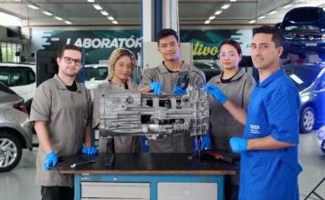 SENAI/SC lidera ranking nacional de excelência industrial Uma foto mostra cinco pessoas, representando estudantes e um instrutor, reunidas em um ambiente de oficina mecânica. Quatro pessoas mais jovens usam batas de trabalho e luvas azuis, enquanto o instrutor veste uma camisa azul-royal com o patch "SENAI Educação Profissional". Eles estão ao redor de um grande componente mecânico de veículo, provavelmente um bloco de motor, apoiado em um carrinho de ferramentas de metal. Um estudante segura um dispositivo de diagnóstico eletrônico, que está conectado por fios ao componente. Atrás deles, um grande letreiro na parede lê-se "LABORATÓRIO", e vários carros são visíveis, incluindo um em um elevador. Esta cena demonstra a metodologia "mão na massa", descrita como um diferencial para o SENAI/SC no texto, que é uma instituição de referência em educação profissional há mais de 70 anos. O treinamento prático neste ambiente faz parte de como o SENAI/SC prepara profissionais qualificados para o setor industrial, contribuindo para uma taxa de empregabilidade superior a 90%.
