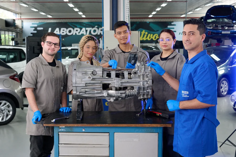 Uma foto mostra cinco pessoas, representando estudantes e um instrutor, reunidas em um ambiente de oficina mecânica. Quatro pessoas mais jovens usam batas de trabalho e luvas azuis, enquanto o instrutor veste uma camisa azul-royal com o patch 