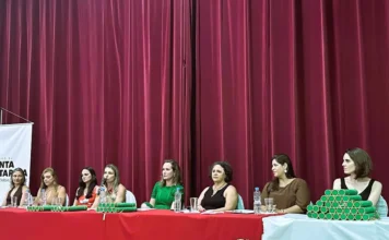 SES forma 70 especialistas para reforçar saúde em SC A imagem mostra a mesa de honra da formatura do programa FEPAPS, composta por oito mulheres sentadas atrás de uma mesa com toalha vermelha. Sobre a mesa, veem-se pilhas de canudos de diplomas na cor verde e copos de água. Uma das integrantes da mesa fala ao microfone, enquanto as demais acompanham a solenidade. Ao fundo, há uma cortina bordô e, em primeiro plano, um arranjo de flores brancas.