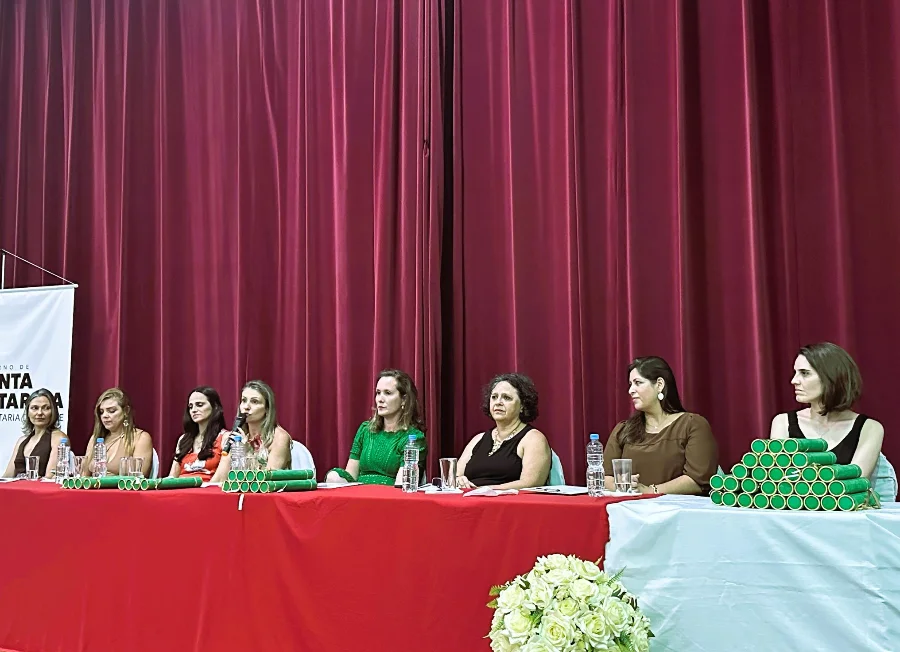 SES forma 70 especialistas para reforçar saúde em SC A imagem mostra a mesa de honra da formatura do programa FEPAPS, composta por oito mulheres sentadas atrás de uma mesa com toalha vermelha. Sobre a mesa, veem-se pilhas de canudos de diplomas na cor verde e copos de água. Uma das integrantes da mesa fala ao microfone, enquanto as demais acompanham a solenidade. Ao fundo, há uma cortina bordô e, em primeiro plano, um arranjo de flores brancas.