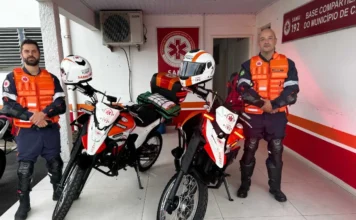 Samu inicia operação de motolâncias em Criciúma Dois socorristas do Samu de Criciúma, trajando uniformes azuis com coletes de proteção laranjas, posam ao lado de duas motolâncias brancas e laranjas estacionadas em frente à base do serviço. As motocicletas estão devidamente equipadas com capacetes sobre o guidão e baús traseiros que transportam kits de primeiros socorros e equipamentos médicos. Ao fundo, é possível ver a placa de identificação da "Base Compartilhada do Município de Criciúma".