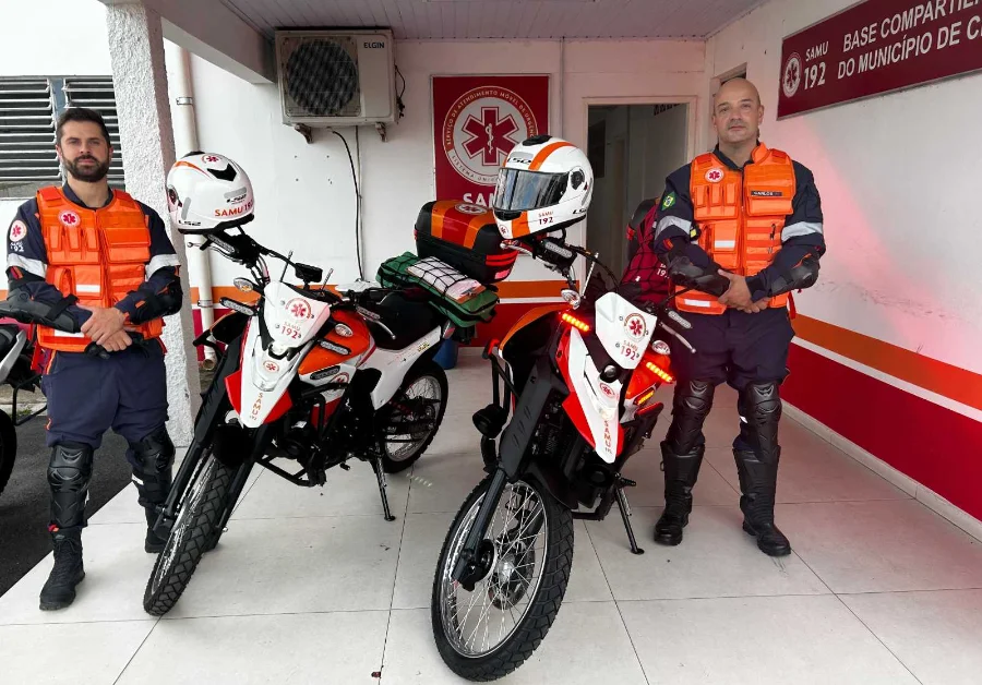 Samu inicia operação de motolâncias em Criciúma Dois socorristas do Samu de Criciúma, trajando uniformes azuis com coletes de proteção laranjas, posam ao lado de duas motolâncias brancas e laranjas estacionadas em frente à base do serviço. As motocicletas estão devidamente equipadas com capacetes sobre o guidão e baús traseiros que transportam kits de primeiros socorros e equipamentos médicos. Ao fundo, é possível ver a placa de identificação da