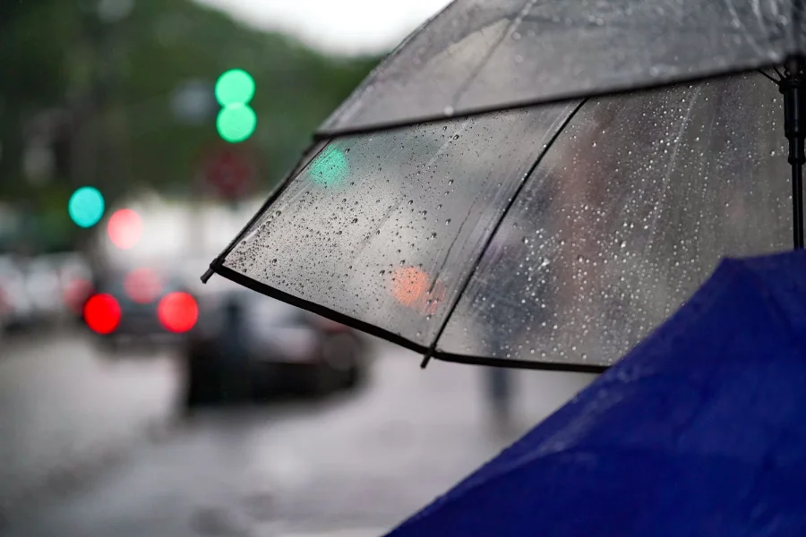 Uma foto de close-up foca em dois guarda-chuvas sob chuva em uma rua urbana desfocada. O guarda-chuva transparente está no primeiro plano, inclinado, com sua superfície coberta por inúmeras gotas de água cristalinas e escorrendo. Os raios e a borda preta do guarda-chuva transparente são nítidos. No canto inferior direito, a borda desfocada de um guarda-chuva azul cobalto profundo é visível. O fundo é uma cena de rua urbana altamente desfocada (bokeh), onde luzes de semáforo verdes e lanternas traseiras de carros vermelhas criam borrões coloridos e atmosféricos contra a paisagem urbana escura e úmida. A profundidade de campo é muito rasa, acentuando a sensação de chuva e isolamento.