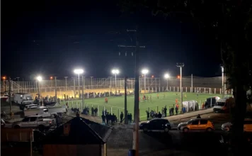 Santa Rosa do Sul inicia Campeonato de Society dia 9 Uma vista noturna em plano médio e ligeiramente elevada da arena esportiva do Parque Municipal da Polvilhana, em Santa Rosa do Sul. O campo de futebol society, com grama sintética verde, está totalmente iluminado por potentes refletores, e um jogo está em andamento. Jogadores uniformizados de vermelho e verde/amarelo são visíveis no campo, movimentando-se ao redor da bola. Atrás de uma cerca metálica que circunda o gramado, um grande número de espectadores está em pé, acompanhando a partida. A área ao redor do complexo esportivo está movimentada, com diversos carros, picapes e vans estacionados. Em primeiro plano, vê-se o telhado de um quiosque de madeira e postes de iluminação de rua. A imagem captura a atmosfera vibrante e o movimento de uma noite de competição do Campeonato Society 2026.