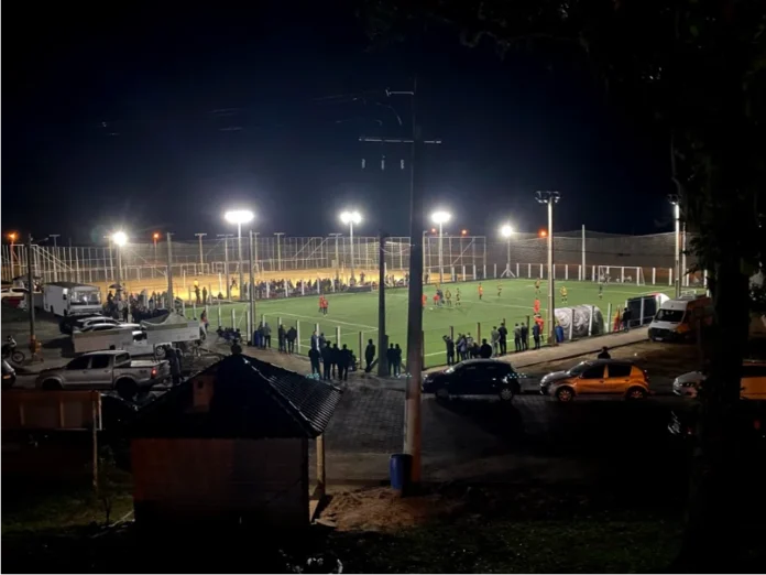 Santa Rosa do Sul inicia Campeonato de Society dia 9 Uma vista noturna em plano médio e ligeiramente elevada da arena esportiva do Parque Municipal da Polvilhana, em Santa Rosa do Sul. O campo de futebol society, com grama sintética verde, está totalmente iluminado por potentes refletores, e um jogo está em andamento. Jogadores uniformizados de vermelho e verde/amarelo são visíveis no campo, movimentando-se ao redor da bola. Atrás de uma cerca metálica que circunda o gramado, um grande número de espectadores está em pé, acompanhando a partida. A área ao redor do complexo esportivo está movimentada, com diversos carros, picapes e vans estacionados. Em primeiro plano, vê-se o telhado de um quiosque de madeira e postes de iluminação de rua. A imagem captura a atmosfera vibrante e o movimento de uma noite de competição do Campeonato Society 2026.