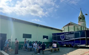 Santa Rosa do Sul realiza mutirão de castração de animais Mutirão de castração móvel do programa Pet Levado a Sério em Santa Rosa do Sul. Tutores de animais de estimação sentados ao ar livre, aguardando atendimento em frente a um trailer médico veterinário e a uma igreja com torre branca e verde-claro.