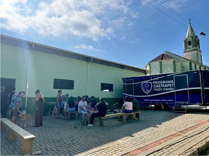Santa Rosa do Sul realiza mutirão de castração de animais Mutirão de castração móvel do programa Pet Levado a Sério em Santa Rosa do Sul. Tutores de animais de estimação sentados ao ar livre, aguardando atendimento em frente a um trailer médico veterinário e a uma igreja com torre branca e verde-claro.