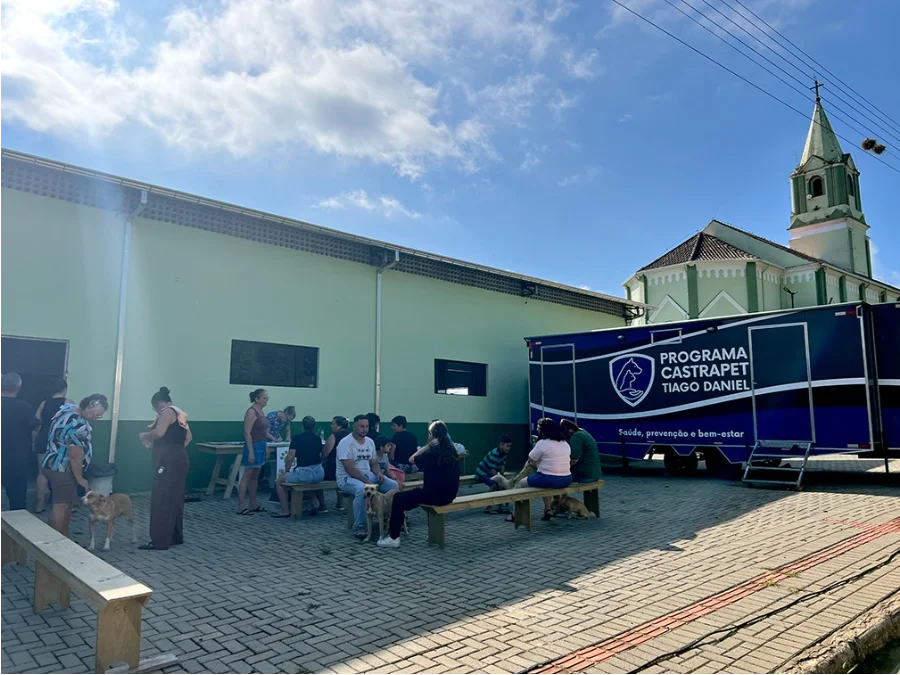 Santa Rosa do Sul realiza mutirão de castração de animais Mutirão de castração móvel do programa Pet Levado a Sério em Santa Rosa do Sul. Tutores de animais de estimação sentados ao ar livre, aguardando atendimento em frente a um trailer médico veterinário e a uma igreja com torre branca e verde-claro.