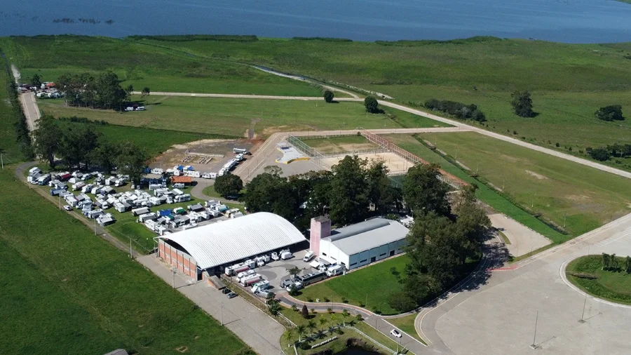 Vista aérea do 1º Encontro de Motorhomes 2026 no Parque Municipal da Polvilhana, em Santa Rosa do Sul, Santa Catarina. A imagem mostra dezenas de veículos de recreação do Grupo Rodamundo estacionados entre os pavilhões do parque e as margens da Lagoa do Sombrio.