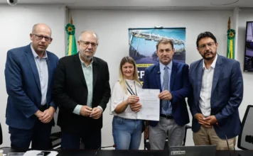 Secretário de Logística falta à CPI após liminar judicial Reunião da CPI das Rodovias na Assembleia Legislativa do Rio Grande do Sul (ALRS). Cinco pessoas, incluindo o deputado estadual Miguel Rossetto (com placa de identificação à direita), recebem um documento oficial. No fundo, bandeiras do RS e um pôster da 'Comissão Parlamentar de Inquérito' com imagem de pedágio. Cena focada na entrega de requerimentos ou mandados relacionados à investigação de contratos de concessão rodoviária.