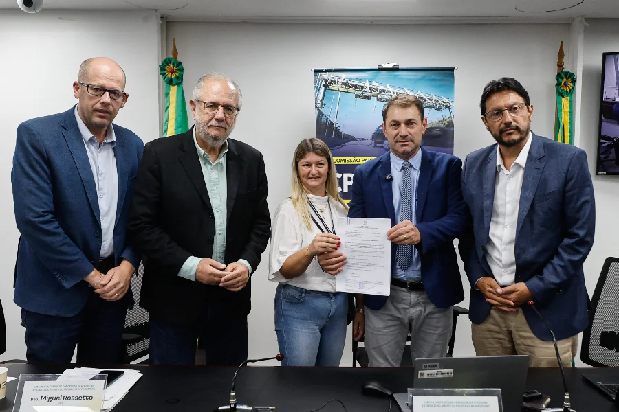 Reunião da CPI das Rodovias na Assembleia Legislativa do Rio Grande do Sul (ALRS). Cinco pessoas, incluindo o deputado estadual Miguel Rossetto (com placa de identificação à direita), recebem um documento oficial. No fundo, bandeiras do RS e um pôster da 'Comissão Parlamentar de Inquérito' com imagem de pedágio. Cena focada na entrega de requerimentos ou mandados relacionados à investigação de contratos de concessão rodoviária.