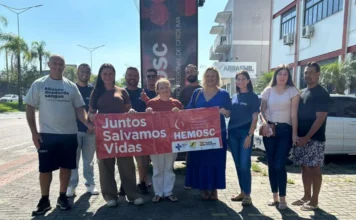 Servidores do SAMAE de Araranguá doam sangue no Hemosc Servidores do SAMAE de Araranguá participando de ação coletiva de doação de sangue em frente ao Hemosc Criciúma, segurando um banner vermelho com a inscrição 'Juntos Salvamos Vidas'.