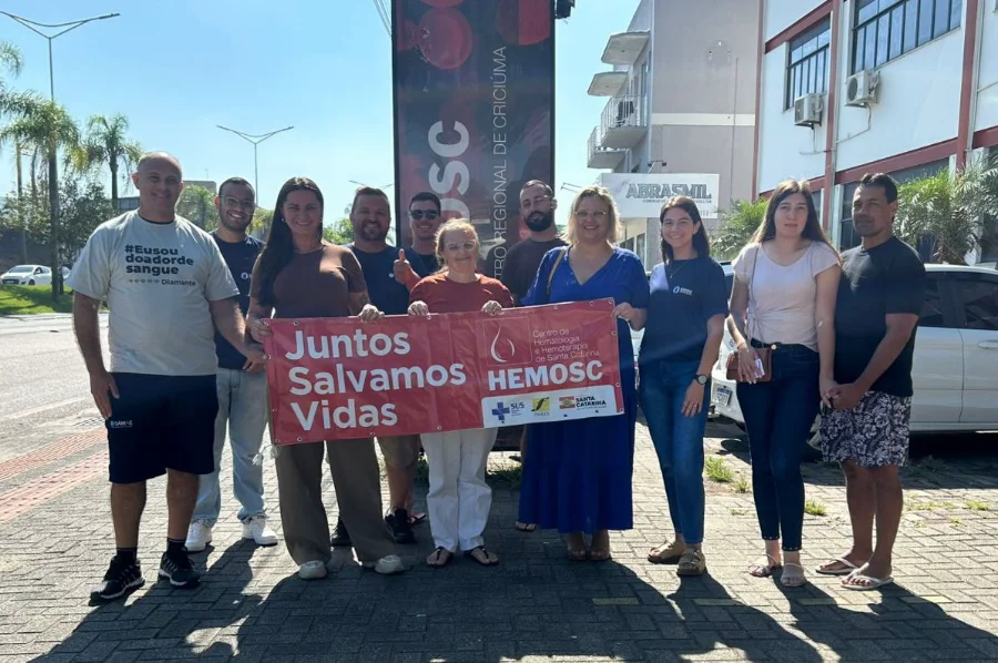 Servidores do SAMAE de Araranguá doam sangue no Hemosc Servidores do SAMAE de Araranguá participando de ação coletiva de doação de sangue em frente ao Hemosc Criciúma, segurando um banner vermelho com a inscrição 'Juntos Salvamos Vidas'.