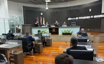 Sessão em Araranguá destaca pontes e programa de reciclagem Fotografia em perspectiva do plenário da Câmara de Vereadores de Araranguá durante uma sessão oficial. Em primeiro plano, vê-se a parte posterior da cabeça de um parlamentar, papéis e um copo de água sobre a bancada de madeira. O salão possui piso de madeira polida e diversas fileiras de mesas ocupadas por vereadores em trajes formais. Ao fundo, em um nível elevado, quatro autoridades compõem a mesa diretora à frente de uma parede ripada escura com o letreiro em destaque "CÂMARA DE VEREADORES DE ARARANGUÁ". Bandeiras oficiais e arranjos de flores brancas completam a composição do cenário legislativo.
