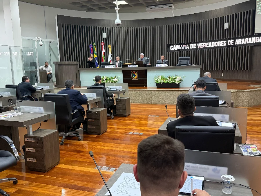Fotografia em perspectiva do plenário da Câmara de Vereadores de Araranguá durante uma sessão oficial. Em primeiro plano, vê-se a parte posterior da cabeça de um parlamentar, papéis e um copo de água sobre a bancada de madeira. O salão possui piso de madeira polida e diversas fileiras de mesas ocupadas por vereadores em trajes formais. Ao fundo, em um nível elevado, quatro autoridades compõem a mesa diretora à frente de uma parede ripada escura com o letreiro em destaque 