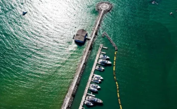 Economia do mar impulsiona mercado de trabalho em SC Vista aérea de uma infraestrutura marítima no litoral de Santa Catarina, exemplificando a Economia do Mar. Um longo quebra-mar de pedras se estende para as águas verdes-esmeralda, terminando em uma plataforma circular com uma rosa dos ventos no design. Paralela ao quebra-mar, uma doca flutuante abriga uma fileira de iates e barcos de lazer de diversos tamanhos, simbolizando os setores de construção de embarcações e serviços de marina. Um edifício com telhado escuro, possivelmente um hotel ou restaurante, está localizado próximo à doca. No mar mais claro à esquerda, um pequeno barco isolado é visível, enquanto uma linha de boias amarelas flutua à direita da doca.