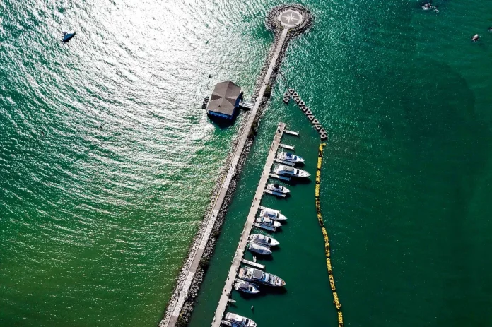 Setor marinho responde por 13% das novas vagas em SC Vista aérea de uma infraestrutura marítima no litoral de Santa Catarina, exemplificando a Economia do Mar. Um longo quebra-mar de pedras se estende para as águas verdes-esmeralda, terminando em uma plataforma circular com uma rosa dos ventos no design. Paralela ao quebra-mar, uma doca flutuante abriga uma fileira de iates e barcos de lazer de diversos tamanhos, simbolizando os setores de construção de embarcações e serviços de marina. Um edifício com telhado escuro, possivelmente um hotel ou restaurante, está localizado próximo à doca. No mar mais claro à esquerda, um pequeno barco isolado é visível, enquanto uma linha de boias amarelas flutua à direita da doca.