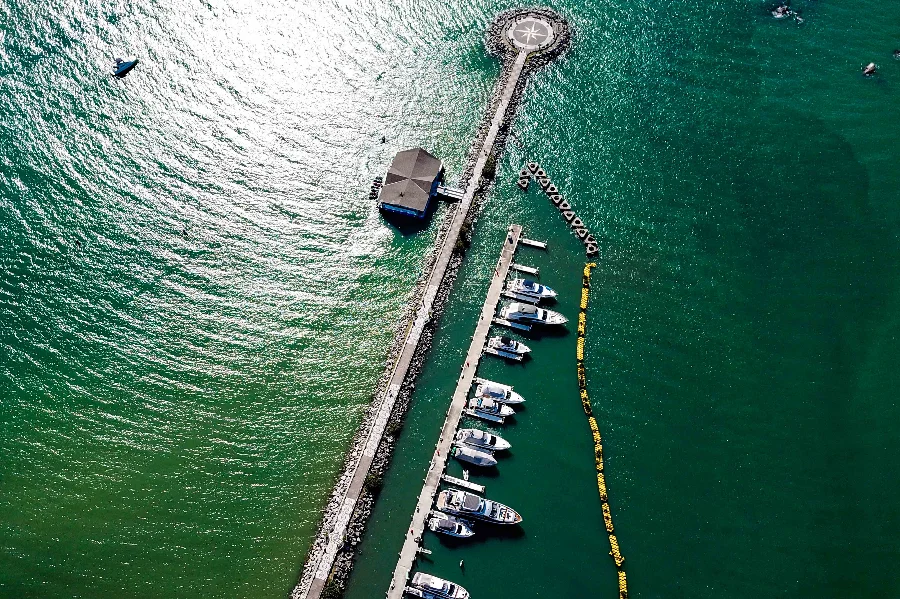Vista aérea de uma infraestrutura marítima no litoral de Santa Catarina, exemplificando a Economia do Mar. Um longo quebra-mar de pedras se estende para as águas verdes-esmeralda, terminando em uma plataforma circular com uma rosa dos ventos no design. Paralela ao quebra-mar, uma doca flutuante abriga uma fileira de iates e barcos de lazer de diversos tamanhos, simbolizando os setores de construção de embarcações e serviços de marina. Um edifício com telhado escuro, possivelmente um hotel ou restaurante, está localizado próximo à doca. No mar mais claro à esquerda, um pequeno barco isolado é visível, enquanto uma linha de boias amarelas flutua à direita da doca.