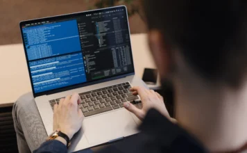 Siderópolis abre vagas para cursos gratuitos em tecnologia Fotografia em plano fechado, vista pelas costas de um homem sentado que utiliza um notebook apoiado no colo. Na tela do computador, observam-se linhas de códigos de programação em cores azul e branco sobre um fundo escuro.