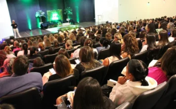 Simpósio na Unesc debate autismo além da infância Público lota auditório em Criciúma durante palestra do 7º Simpósio LAND da Unesc sobre autismo. A imagem mostra as pessoas sentadas acompanhando a apresentação no palco decorado com iluminação verde e plantas.