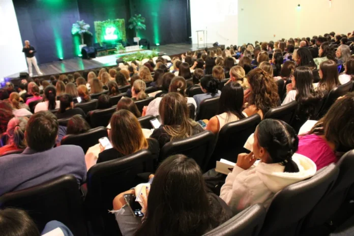 Simpósio na Unesc debate autismo além da infância Público lota auditório em Criciúma durante palestra do 7º Simpósio LAND da Unesc sobre autismo. A imagem mostra as pessoas sentadas acompanhando a apresentação no palco decorado com iluminação verde e plantas.