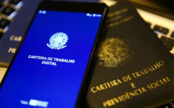 Sine de Morro da Fumaça abre 224 novas vagas de emprego Fotografia em primeiro plano e ângulo levemente inclinado mostra um smartphone com a tela azul brilhante exibindo o logotipo do Governo Federal e o título "Carteira de Trabalho Digital". Ao lado do aparelho, repousa uma Carteira de Trabalho física tradicional, de capa preta e letras douradas. Os itens estão sobre o que parece ser o teclado de um computador, simbolizando o processo de busca por emprego e a documentação necessária para as 224 vagas abertas no Sine de Morro da Fumaça.