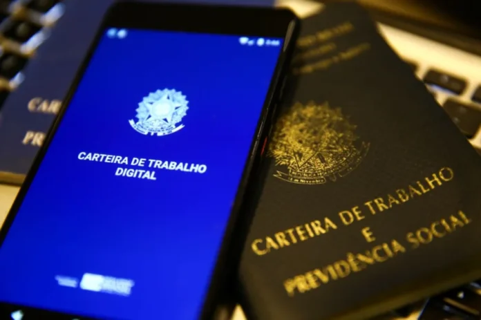 Sine de Morro da Fumaça abre 224 novas vagas de emprego Fotografia em primeiro plano e ângulo levemente inclinado mostra um smartphone com a tela azul brilhante exibindo o logotipo do Governo Federal e o título "Carteira de Trabalho Digital". Ao lado do aparelho, repousa uma Carteira de Trabalho física tradicional, de capa preta e letras douradas. Os itens estão sobre o que parece ser o teclado de um computador, simbolizando o processo de busca por emprego e a documentação necessária para as 224 vagas abertas no Sine de Morro da Fumaça.