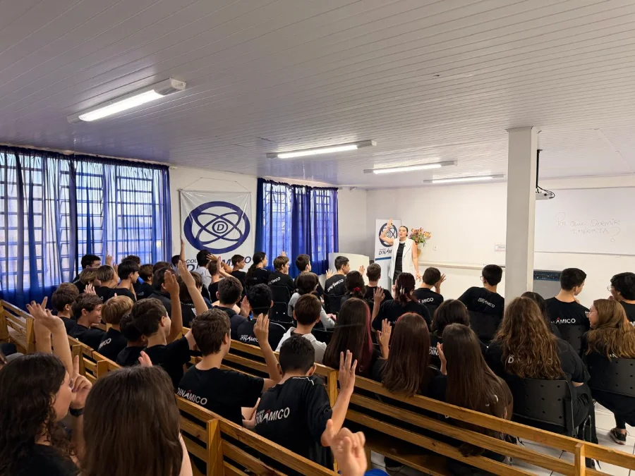 Foto em plano geral mostrando um auditório repleto de estudantes do Colégio Dinâmico, todos vestindo camisetas pretas e sentados em bancos de madeira, com a maioria deles levantando as mãos em um gesto de interação. No palco à direita, uma palestrante feminina, de camisa branca e calça preta, está de pé e gesticulando com um braço levantado enquanto fala. Atrás dela, há uma mesa com flores, um quadro branco com escritas parciais e um banner da escola com um logotipo de átomo. O ambiente tem janelas com cortinas azuis ao fundo e luminárias fluorescentes no teto.