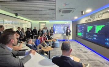 Suíços buscam parcerias comerciais em Santa Catarina Fotografia em plano aberto mostra o interior de um moderno lounge de conferência. Um grupo de cerca de vinte pessoas, vestindo trajes de negócios, está sentado em dois níveis de assentos de estilo arquibancada, de frente para um grande video wall que cobre quase toda a parede do fundo. As telas do video wall exibem várias visualizações de dados, incluindo gráficos de barras, gráficos de linhas e mapas de calor, tudo em tons de verde e azul. Um homem de terno cinza está em pé no centro-direita, gesticulando para as telas enquanto faz uma apresentação. Acima do video wall, há um logotipo em relevo e o texto "DLAB OBSERVATÓRIO FIESC". O ambiente tem teto acústico e paredes de vidro ao fundo. À esquerda, outro grupo de pessoas está em pé assistindo à apresentação. A iluminação é sutil e moderna, com frestas de LED azul.