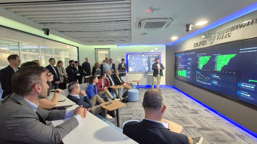 Fotografia em plano aberto mostra o interior de um moderno lounge de conferência. Um grupo de cerca de vinte pessoas, vestindo trajes de negócios, está sentado em dois níveis de assentos de estilo arquibancada, de frente para um grande video wall que cobre quase toda a parede do fundo. As telas do video wall exibem várias visualizações de dados, incluindo gráficos de barras, gráficos de linhas e mapas de calor, tudo em tons de verde e azul. Um homem de terno cinza está em pé no centro-direita, gesticulando para as telas enquanto faz uma apresentação. Acima do video wall, há um logotipo em relevo e o texto 