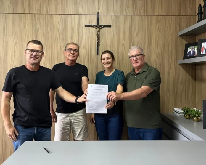 Timbé do Sul assina ordem para obra de farmácia pública Prefeito Vilmar Maffiolette e a Secretária de Saúde de Timbé do Sul, Sabrina Moro Pizzolo, ladeados por outras autoridades municipais, seguram o documento de ordem de serviço assinado para o início das obras da nova farmácia pública municipal, anexa ao Posto Central. Foto posada em escritório de prefeitura.