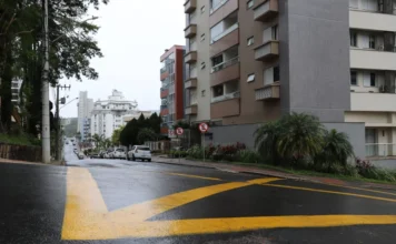 Trecho da rua Lauro Müller terá mão única em Criciúma Vista de uma rua urbana íngreme e molhada em um dia nublado. Faixas de sinalização amarelas brilhantes são proeminentes no asfalto molhado no primeiro plano, indicando direções de tráfego. Prédios de apartamentos de vários andares revestem o lado direito da rua, e uma colina arborizada está no lado esquerdo. Carros estão estacionados ao longo da rua subindo a colina na distância.