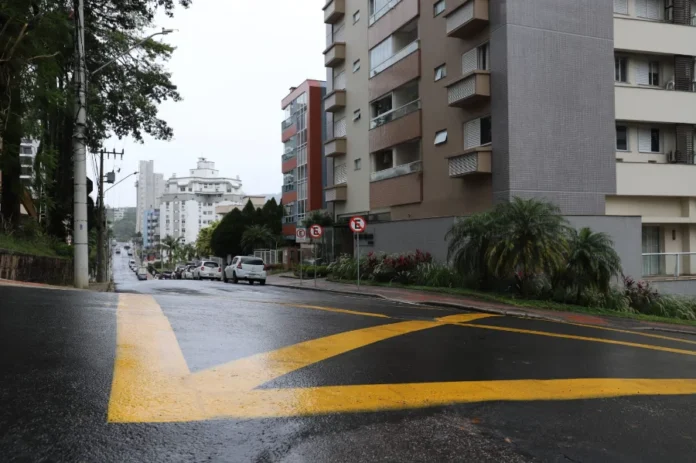 Trecho da rua Lauro Müller terá mão única em Criciúma Vista de uma rua urbana íngreme e molhada em um dia nublado. Faixas de sinalização amarelas brilhantes são proeminentes no asfalto molhado no primeiro plano, indicando direções de tráfego. Prédios de apartamentos de vários andares revestem o lado direito da rua, e uma colina arborizada está no lado esquerdo. Carros estão estacionados ao longo da rua subindo a colina na distância.