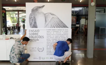 Unesc abre exposição “Ensaio sobre Coleções” nesta quarta Ajoelhados no saguão da Unesc, dois funcionários fazem a instalação do grande painel de divulgação para a exposição "Ensaio sobre Coleções", da artista visual Elisa Corrêa. O painel exibe, no topo, uma detalhada ilustração em grafite de um pássaro, e abaixo, o texto explicativo sobre a 152ª edição do projeto Espaço Cultural Unesc "Toque de Arte" e a pesquisa da artista com elementos da natureza. A cena detalha o processo de montagem e o texto do banner.