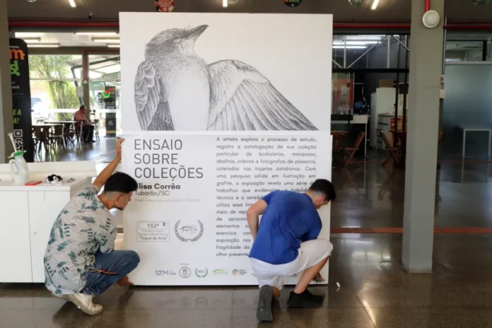 Unesc abre exposição Ensaio sobre Coleções nesta quarta Ajoelhados no saguão da Unesc, dois funcionários fazem a instalação do grande painel de divulgação para a exposição "Ensaio sobre Coleções", da artista visual Elisa Corrêa. O painel exibe, no topo, uma detalhada ilustração em grafite de um pássaro, e abaixo, o texto explicativo sobre a 152ª edição do projeto Espaço Cultural Unesc "Toque de Arte" e a pesquisa da artista com elementos da natureza. A cena detalha o processo de montagem e o texto do banner.