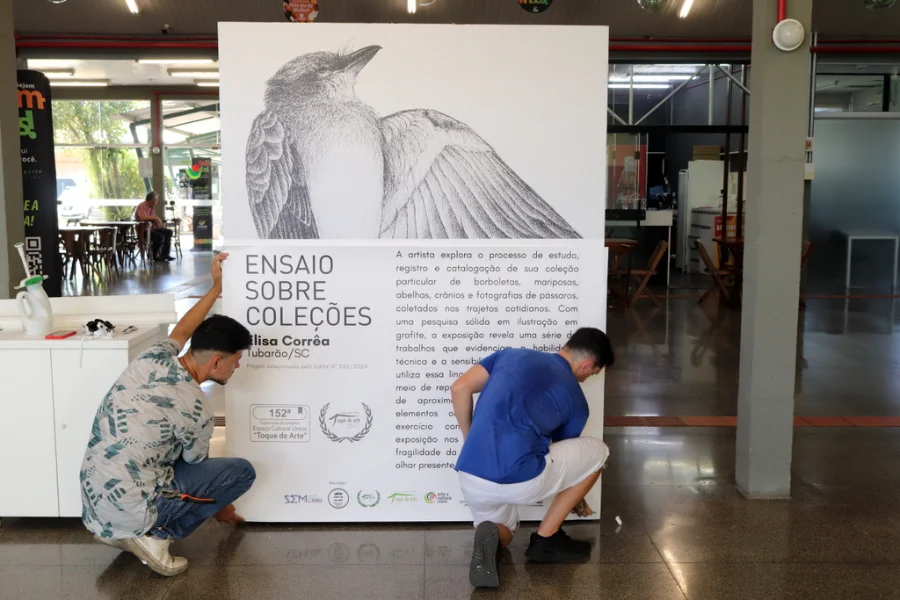 Ajoelhados no saguão da Unesc, dois funcionários fazem a instalação do grande painel de divulgação para a exposição 