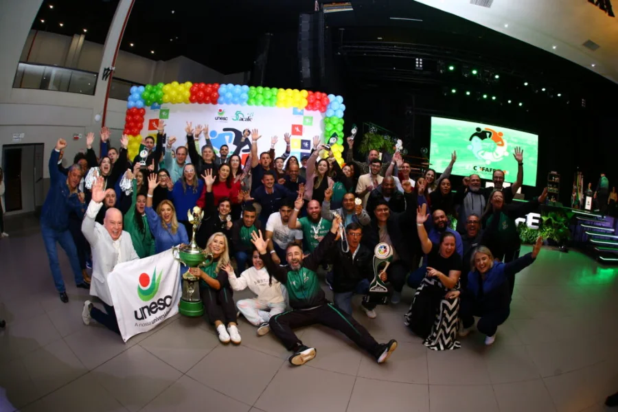 Delegação campeã da Unesc em celebração com o troféu geral da Cofafe (Acafe) no palco. Professores e funcionários seguram medalhas e uma bandeira da instituição, diante de um banner colorido e telão de LED.
