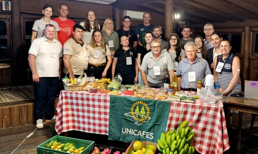 Grupo de participantes do curso de formação para dirigentes cooperativistas da Unesc reunidos atrás de uma mesa com produtos da agricultura familiar, como bananas e pitayas, em Jaguaruna. No centro, destaca-se a bandeira da Unicafes.