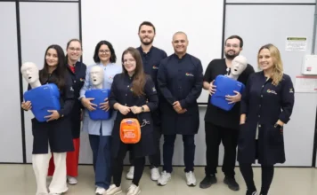 Unesc capacita profissionais em primeiros socorros "Um grupo de oito profissionais de saúde das Clínicas Integradas Unesc, composto por cinco mulheres e três homens, posa sorrindo para a câmera em uma sala de treinamento, após a conclusão de uma capacitação em primeiros socorros e ressuscitação. Eles estão vestindo jalecos médicos (na sua maioria azuis escuros com o logotipo da instituição) e uniformes de médico. Três pessoas seguram manequins de treinamento de RCP (ressuscitação cardiopulmonar) azuis, e uma mulher no centro segura um estojo de DEA (Desfibrilador Externo Automático) laranja. O fundo mostra um quadro branco em branco e sinalização de parede em português, como 'AR CONDICIONADO' e 'Extintor'. O piso é de linóleo. A imagem ilustra a equipe preparada para lidar com possíveis emergências médicas na clínica."