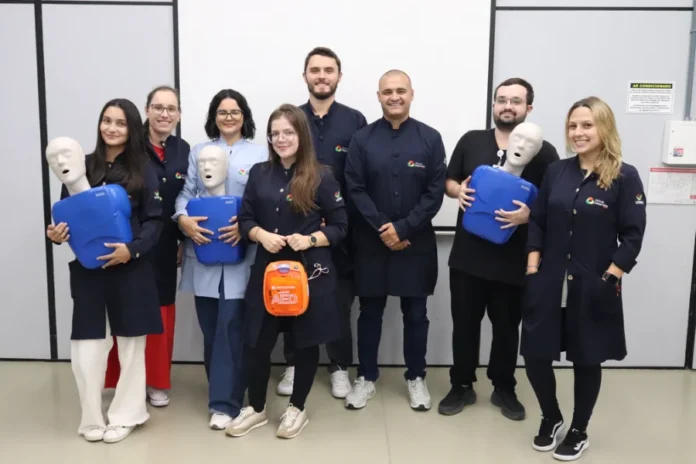 Unesc capacita profissionais em primeiros socorros "Um grupo de oito profissionais de saúde das Clínicas Integradas Unesc, composto por cinco mulheres e três homens, posa sorrindo para a câmera em uma sala de treinamento, após a conclusão de uma capacitação em primeiros socorros e ressuscitação. Eles estão vestindo jalecos médicos (na sua maioria azuis escuros com o logotipo da instituição) e uniformes de médico. Três pessoas seguram manequins de treinamento de RCP (ressuscitação cardiopulmonar) azuis, e uma mulher no centro segura um estojo de DEA (Desfibrilador Externo Automático) laranja. O fundo mostra um quadro branco em branco e sinalização de parede em português, como 'AR CONDICIONADO' e 'Extintor'. O piso é de linóleo. A imagem ilustra a equipe preparada para lidar com possíveis emergências médicas na clínica."