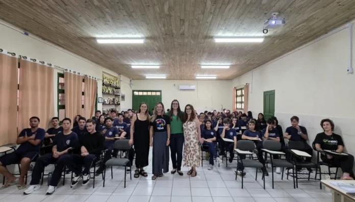 Unesc debate combate à violência contra a mulher em escola Uma foto de grupo em uma sala de aula com teto de madeira. No centro, a professora da Unesc Vanessa Iribarrem Avena Miranda, acompanhada por outras educadoras e mestrandas da instituição, posa de pé em frente a um grande grupo de estudantes sentados. Os alunos, em uniformes azuis, participaram da ação educativa sobre o combate à violência contra mulheres e meninas.