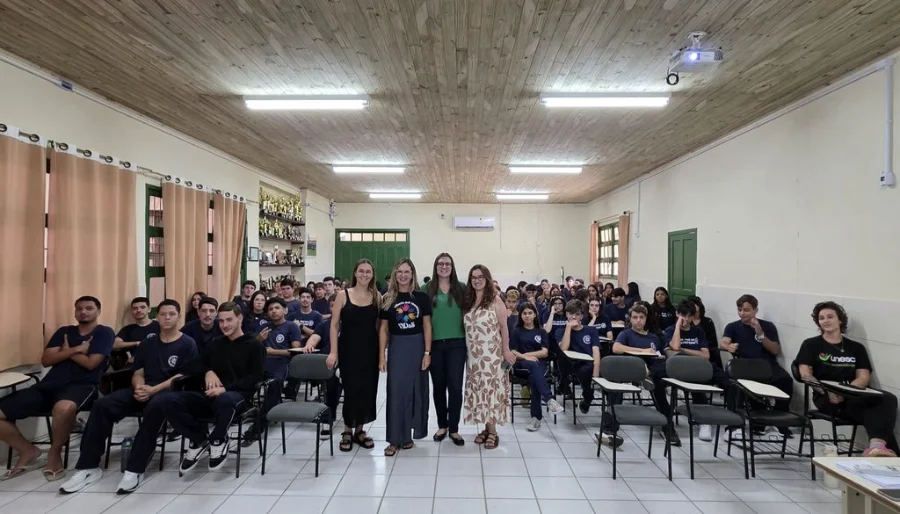 Uma foto de grupo em uma sala de aula com teto de madeira. No centro, a professora da Unesc Vanessa Iribarrem Avena Miranda, acompanhada por outras educadoras e mestrandas da instituição, posa de pé em frente a um grande grupo de estudantes sentados. Os alunos, em uniformes azuis, participaram da ação educativa sobre o combate à violência contra mulheres e meninas.