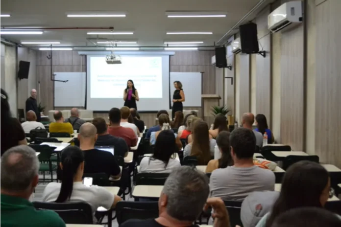 Unesc debate excelência na pós-graduação em Aula Magna Foto de auditório na Unesc com público assistindo a uma apresentação. No palco, a coordenadora da Capes, Verônica Calado, fala ao microfone. No telão, informações sobre a Aula Magna do PPGCEM (Ciência e Engenharia de Materiais) Unesc.