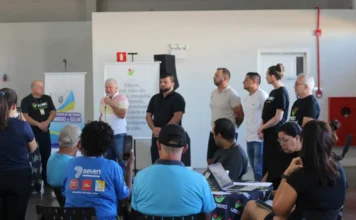 Unesc e Arroio do Silva planejam desenvolvimento para 10 anos Fotografia de um palestrante idoso, com uma camisa polo colorida (amarela, branca e azul) e microfone na mão, falando para uma plateia atenta durante o Workshop da Unesc sobre o Plano de Desenvolvimento Econômico de Balneário Arroio do Silva. Ele está posicionado em frente a um banner do município de Balneário Arroio do Silva e ladeado por outras pessoas de pé. No primeiro plano, membros da plateia assistem, incluindo um homem de costas vestindo uma camisa azul da marca 'seven' e uma mulher tirando uma foto com seu smartphone. Uma mesa com uma toalha com o logo da Unesc e um laptop aberto estão visíveis no primeiro plano. O local é o Centro Multiuso Esther Pacheco Mucillo.