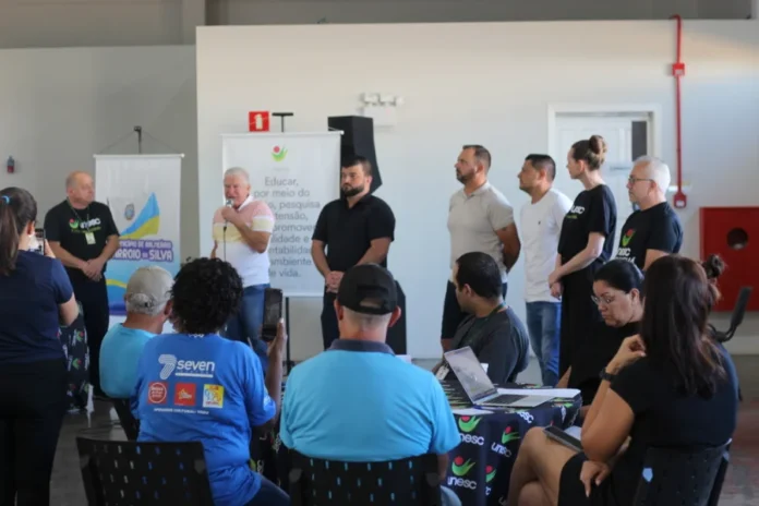 Unesc e Arroio do Silva planejam desenvolvimento para 10 anos Fotografia de um palestrante idoso, com uma camisa polo colorida (amarela, branca e azul) e microfone na mão, falando para uma plateia atenta durante o Workshop da Unesc sobre o Plano de Desenvolvimento Econômico de Balneário Arroio do Silva. Ele está posicionado em frente a um banner do município de Balneário Arroio do Silva e ladeado por outras pessoas de pé. No primeiro plano, membros da plateia assistem, incluindo um homem de costas vestindo uma camisa azul da marca 'seven' e uma mulher tirando uma foto com seu smartphone. Uma mesa com uma toalha com o logo da Unesc e um laptop aberto estão visíveis no primeiro plano. O local é o Centro Multiuso Esther Pacheco Mucillo.