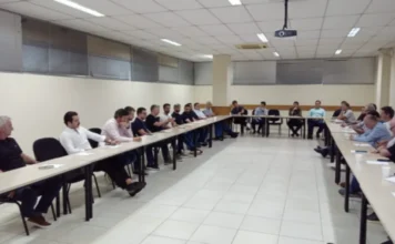 Unesc e Sinduscon reforçam parceria em Criciúma Uma fotografia interna capturada em plano aberto mostra uma reunião formal realizada em uma sala com mesas brancas dispostas em formato de "U". Cerca de 25 homens estão sentados ao redor das mesas, vestindo trajes casuais e executivos, predominantemente camisas polo e camisas sociais. O ambiente possui paredes de cor clara, piso cerâmico branco e iluminação fluorescente embutida no teto. Sobre as mesas, encontram-se copos descartáveis, papéis e materiais de apoio. Ao fundo, é possível observar janelas com persianas cinzas.