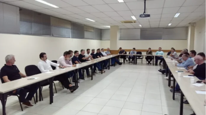 Unesc e Sinduscon reforçam parceria em Criciúma Uma fotografia interna capturada em plano aberto mostra uma reunião formal realizada em uma sala com mesas brancas dispostas em formato de "U". Cerca de 25 homens estão sentados ao redor das mesas, vestindo trajes casuais e executivos, predominantemente camisas polo e camisas sociais. O ambiente possui paredes de cor clara, piso cerâmico branco e iluminação fluorescente embutida no teto. Sobre as mesas, encontram-se copos descartáveis, papéis e materiais de apoio. Ao fundo, é possível observar janelas com persianas cinzas.